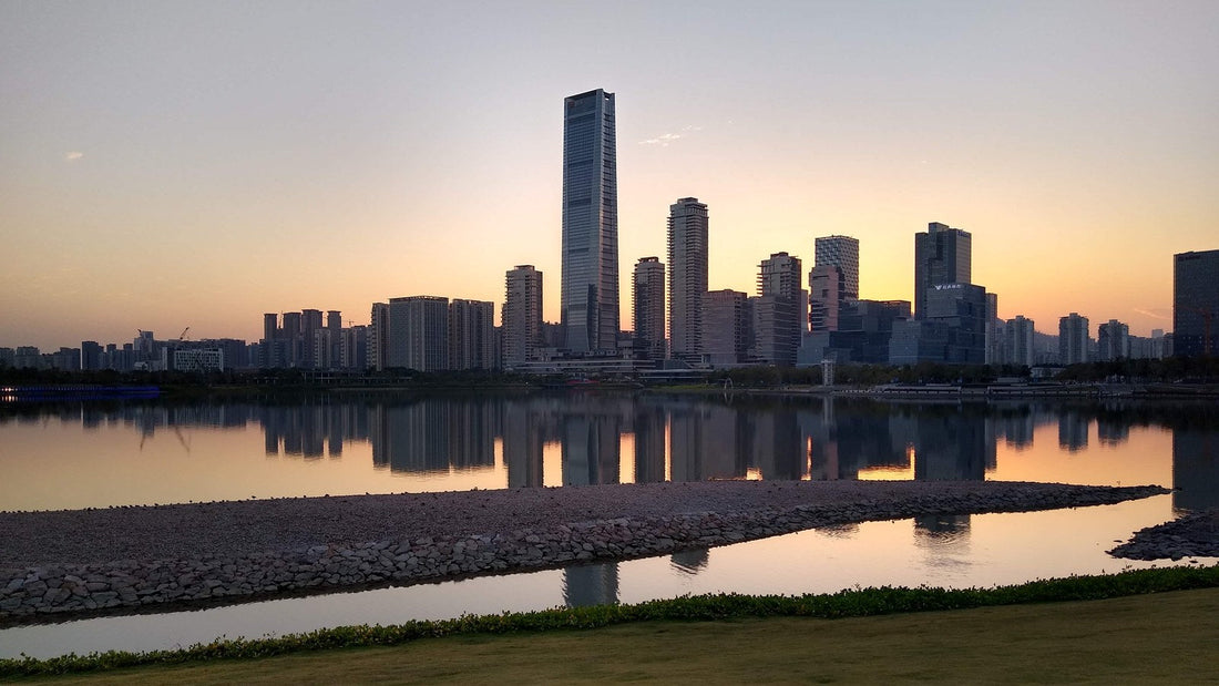 Sunset over the Shenzhen skyline at Shenzhen Bay Park, with tall towers reflected in calm water and a rocky strip and grass in the foreground